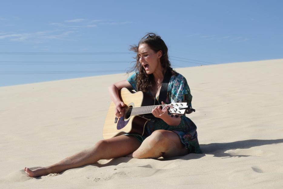 Maddie Grace Newton sittin g on a beach while singing and playing guitar.
