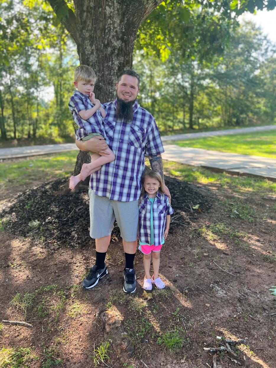 Father Ricky Powell and his two children in matching blue plaid shirts standing in front of a large tree