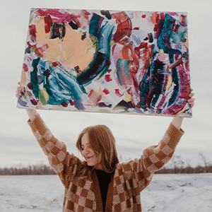 Medora Frei standing outside and holding a painting of hers over her head