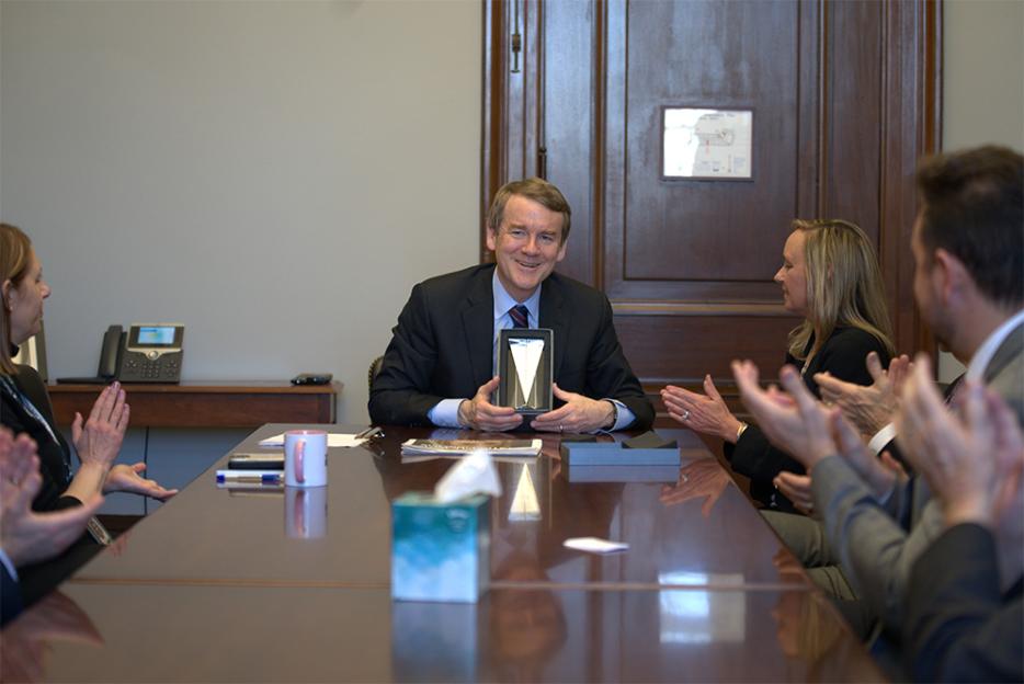 Sen. Michael Bennet (D-CO) holding an award at a conference table while those seated at the table clap.