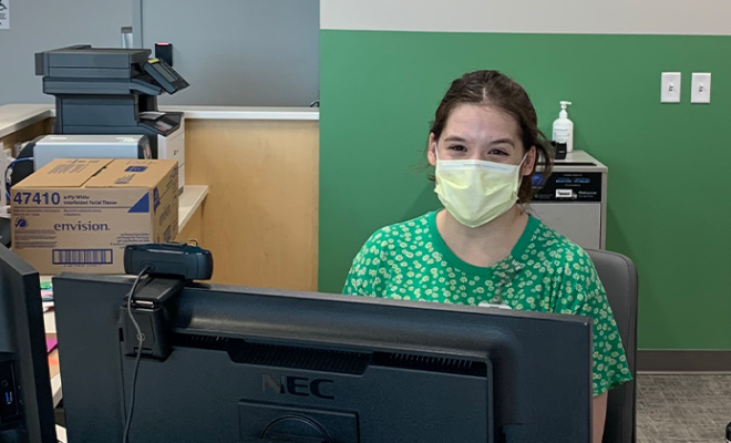 Natalie smiling with her mask on sitting at a desk.