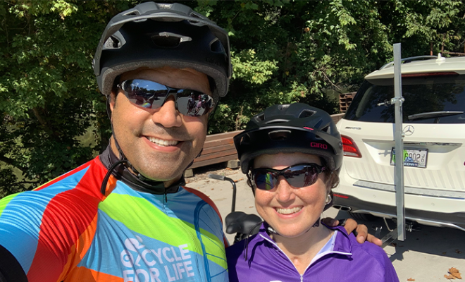 Ray smiling with his wife Rebecca wearing bike helmets and sunglasses.